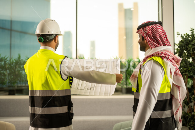 مشاريع إعمارية سعودية وطنية، مفهوم الهندسة والإنشاءات المعمارية، التفاهم والتعاون بين الزملاء، مهندس عربي خليجي سعودي يرتدي سترة العمل وخوذة الحماية البيضاء مع زميله يقرأوا المخططات الهندسية