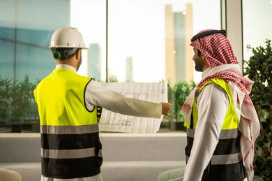 مشاريع إعمارية سعودية وطنية، مفهوم الهندسة والإنشاءات المعمارية، التفاهم والتعاون بين الزملاء، مهندس عربي خليجي سعودي يرتدي سترة العمل وخوذة الحماية البيضاء مع زميله يقرأوا المخططات الهندسية