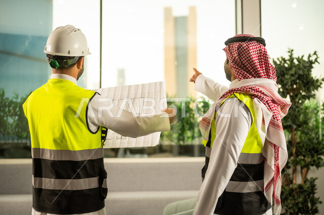 التفاهم والتعاون بين الزملاء، الاشارة الى شيء ما، مهندس عربي خليجي سعودي يرتدي سترة العمل وخوذة الحماية البيضاء مع زميله يقرأوا المخططات الهندسية، مفهوم الهندسة والإنشاءات المعمارية, مشاريع إعمارية سعودية وطنية