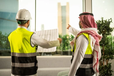 التفاهم والتعاون بين الزملاء، الاشارة الى شيء ما، مهندس عربي خليجي سعودي يرتدي سترة العمل وخوذة الحماية البيضاء مع زميله يقرأوا المخططات الهندسية، مفهوم الهندسة والإنشاءات المعمارية, مشاريع إعمارية سعودية وطنية