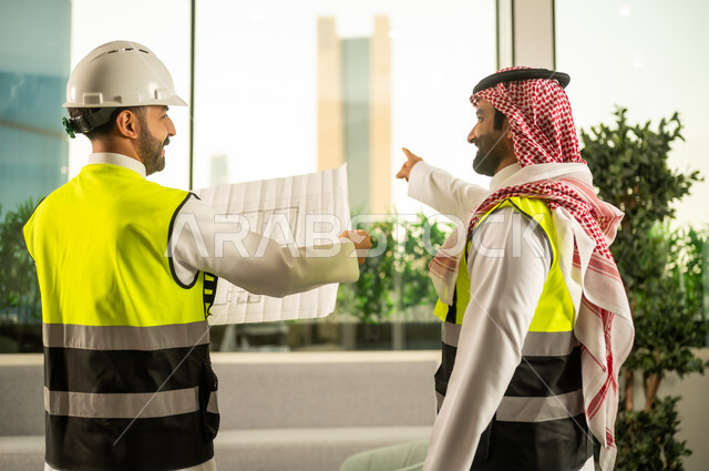 الاشارة الى شيء ما، مهندس عربي خليجي سعودي يرتدي سترة العمل وخوذة الحماية البيضاء مع زميله يقرأوا المخططات الهندسية ، التفاهم والتعاون بين الزملاء، مفهوم الهندسة والإنشاءات المعمارية, مشاريع إعمارية سعودية وطنية