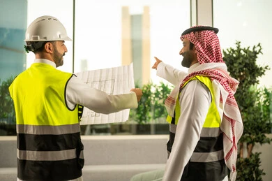الاشارة الى شيء ما، مهندس عربي خليجي سعودي يرتدي سترة العمل وخوذة الحماية البيضاء مع زميله يقرأوا المخططات الهندسية ، التفاهم والتعاون بين الزملاء، مفهوم الهندسة والإنشاءات المعمارية, مشاريع إعمارية سعودية وطنية