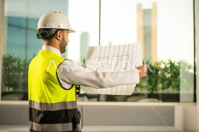 مشاريع إعمارية سعودية وطنية، الاهتمام بالتطور العمراني بسواعد المهندسين السعوديين،  متابعة مخطط المشروع الهندسي وتدقيقه، مفهوم الهندسة المعمارية, مهندس عربي خليجي سعودي يرتدي سترة العمل وخوذة الحماية البيضاء ينجز المهام اليومية في المكتب