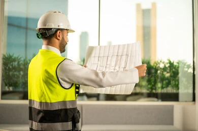 مشاريع إعمارية سعودية وطنية، الاهتمام بالتطور العمراني بسواعد المهندسين السعوديين،  متابعة مخطط المشروع الهندسي وتدقيقه، مفهوم الهندسة المعمارية, مهندس عربي خليجي سعودي يرتدي سترة العمل وخوذة الحماية البيضاء ينجز المهام اليومية في المكتب