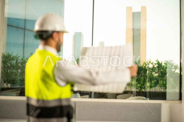 متابعة مخطط المشروع الهندسي وتدقيقه، مفهوم الهندسة المعمارية, مشاريع إعمارية سعودية وطنية، الاهتمام بالتطور العمراني بسواعد المهندسين السعوديين, مهندس عربي خليجي سعودي يرتدي سترة العمل وخوذة الحماية البيضاء ينجز المهام اليومية في المكتب
