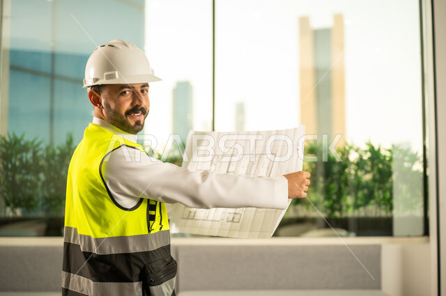 مفهوم الهندسة المعمارية، متابعة مخطط المشروع الهندسي وتدقيقه, مشاريع إعمارية سعودية وطنية، الاهتمام بالتطور العمراني بسواعد المهندسين السعوديين, مهندس عربي خليجي سعودي يرتدي سترة العمل وخوذة الحماية البيضاء ينجز المهام اليومية في المكتب
