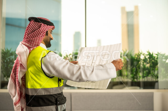 مفهوم دراسة المخطط والمشروع البناء، ادارة المشاريع الهندسية والانشاءات، مهنة ووظيفة هندسية شبابية، مهندس عربي خليجي سعودي يرتدي الثوب التقليدي وسترة الحماية يعمل في القطاع الهندسي في المملكة العربية السعودية يقوم بمراجعة المخطط الهندسي