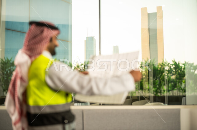 Engineering project management and construction, the concept of studying the plan and construction project, a profession and engineering job for youth, a Saudi Gulf Arab engineer wearing traditional attire and a safety vest working in the engineering sector in the Kingdom of Saudi Arabia reviewing the engineering plan.
