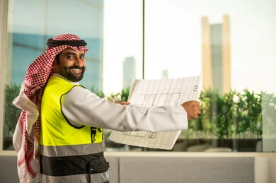 النظر الى الكاميرا بتعابير السرور، ادارة المشاريع الهندسية والانشاءات، مفهوم دراسة المخطط والمشروع البناء، مهنة ووظيفة هندسية شبابية، مهندس عربي خليجي سعودي يرتدي الثوب التقليدي وسترة الحماية يعمل في القطاع الهندسي في المملكة العربية السعودية يقوم بمراجعة المخطط الهندسي