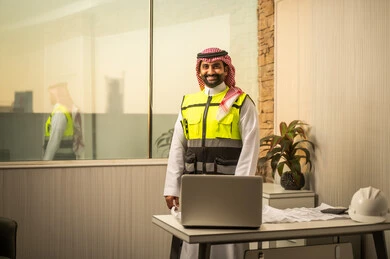 The concept of engineering and construction project management, working in the engineering sector in the Kingdom of Saudi Arabia, a youthful engineering profession and job, a Saudi Gulf Arab engineer wearing traditional attire and a jacket standing inside an engineering office looking at the camera with a joyful expression.