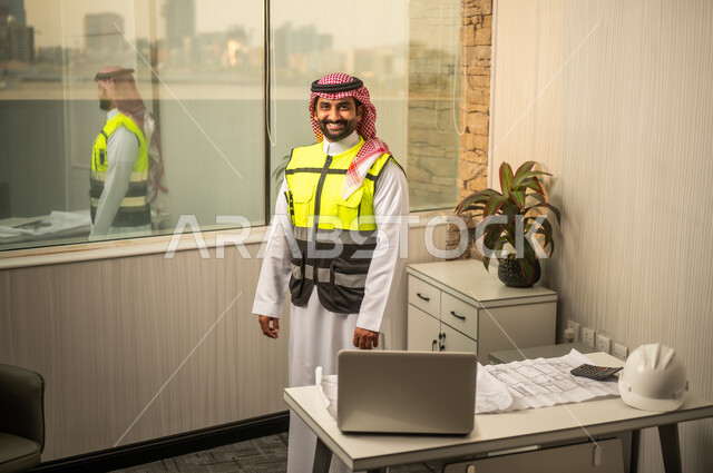 العمل في القطاع الهندسي في المملكة العربية السعودية، مفهوم ادارة المشاريع الهندسية والانشاءات، مهنة ووظيفة هندسية شبابية، مهندس عربي خليجي سعودي يرتدي الثوب التقليدي والسترة يقف داخل مكتب هندسي ينظر الى الكاميرا بتعابير السرور