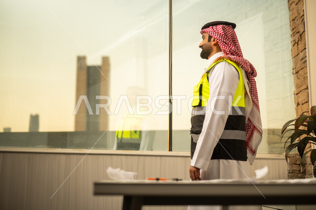 مفهوم التخطيط الاستراتيجي والمتابعة الهندسية والإشراف على مشاريع البناء، صورة من الجانب لمهندس عربي خليجي سعودي يرتدي الشماغ و سترة السلامة المهنية يقف داخل مكتب هندسي ينظر عبر النافذة الزجاجية