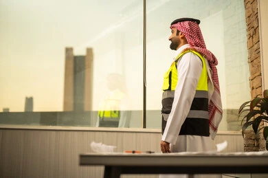 The concept of strategic planning, engineering follow-up, and supervision of construction projects, a side image of a Saudi Gulf Arab engineer wearing a ghutrah and a safety vest standing inside an engineering office looking through the glass window.