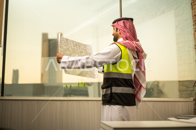 تحليل التصاميم الإنشائية والتأكد من تفاصيل المشروع، النظر للكاميرا بتعابير السرور، مهندس عربي خليجي سعودي يرتدي سترة السلامة و الشماغ يقف داخل مكتب هندسي  يمسك المخطط الورقي ويقوم بمراجعتها، مفهوم التخطيط الهندسي والإشراف على المشاريع