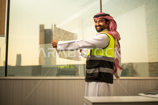 النظر للكاميرا بتعابير السرور، مهندس عربي خليجي سعودي يرتدي سترة السلامة و الشماغ يقف داخل مكتب هندسي  يمسك المخطط الورقي ويقوم بمراجعتها، تحليل التصاميم الإنشائية والتأكد من تفاصيل المشروع، مفهوم التخطيط الهندسي والإشراف على المشاريع