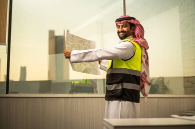 Looking at the camera with expressions of joy, a Saudi Gulf Arab engineer wearing a safety vest and a ghutrah stands inside an engineering office holding a paper plan and reviewing it, analyzing structural designs and ensuring project details, understanding engineering planning and project supervision.