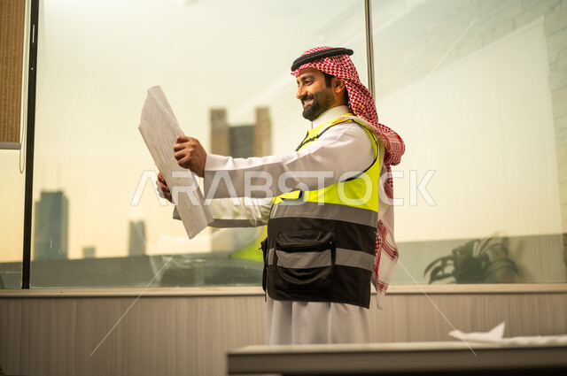 مهندس عربي خليجي سعودي يرتدي سترة السلامة و الشماغ يقف داخل مكتب هندسي  يمسك المخطط الورقي ويقوم بمراجعتها، تحليل التصاميم الإنشائية والتأكد من تفاصيل المشروع، مفهوم التخطيط الهندسي والإشراف على المشاريع
