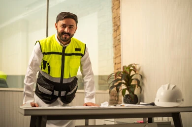 Reviewing paper plans, the concept of engineering management and pre-planning, professional accuracy and engineering work, a Saudi Gulf Arab engineer wearing a thobe and safety vest is reviewing engineering designs in a modern office environment, looking at the camera with a joyful expression.