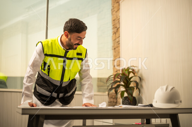 مفهوم الإدارة الهندسية والتخطيط المسبق، الدقة المهنية والعمل الهندسي، مهندس عربي خليجي سعودي يرتدي الثوب و سترة الحماية يعمل على مراجعة التصاميم الهندسية داخل بيئة مكتبية حديثة