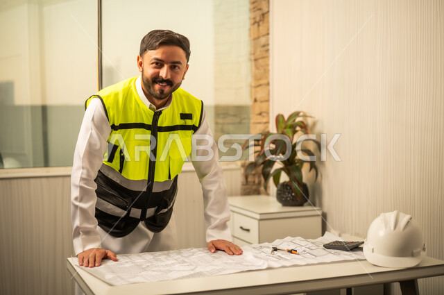 النظر للكاميرا بتعابير السرور،  الدقة المهنية والعمل الهندسي، مهندس عربي خليجي سعودي يرتدي الثوب و سترة الحماية يعمل على مراجعة التصاميم الهندسية داخل بيئة مكتبية حديثة،مفهوم الإدارة الهندسية والتخطيط المسبق