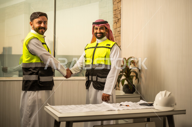 النظر للكاميرا بتعابير السرور، مفهوم التعاون المهني داخل بيئة العمل، التخطيط وإدارة المشاريع الإنشائية، مراجعة المخططات الفنية واتخاذ القرارات، اتفاق عمل قائم على الثقة، مهندس عربي خليجي سعودي يرتدي سترة سلامة و الشماغ يقوم بمصافحة زميلة