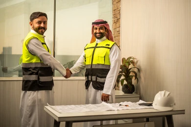 Looking at the camera with expressions of joy, the concept of professional collaboration within the work environment, planning and managing construction projects, reviewing technical drawings and making decisions, a work agreement based on trust, an Arab Gulf Saudi engineer wearing a safety vest and a ghutrah shaking hands with a colleague.