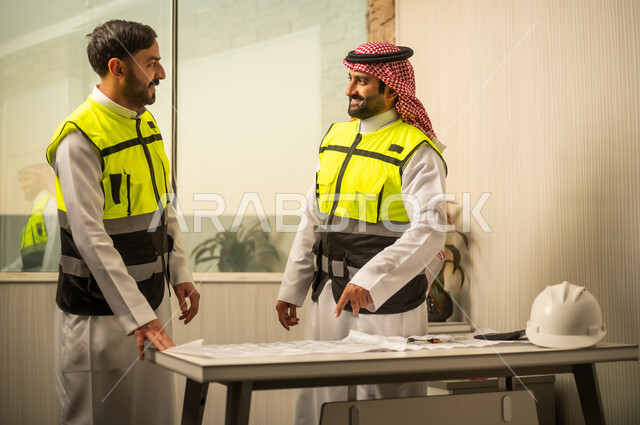 مناقشة مخطط البناء، متابعة سير المشروع، مفهوم التخطيط والتنظيم، العمل الجماعي في بيئة احترافية، مهندس عربي خليجي سعودي يرتدي الشماغ و سترة السلامة يقف برفقة زميلة يشير إلى المخطط الورقي أثناء الشرح