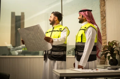 Using paper plans in work, the concept of engineering planning, following up on project details, reviewing the construction plan, a Saudi Gulf Arab engineer wearing a safety vest standing and holding an engineering plan while following the explanation alongside a colleague, a modern work environment in the construction sector, the concept of urban development and project management.