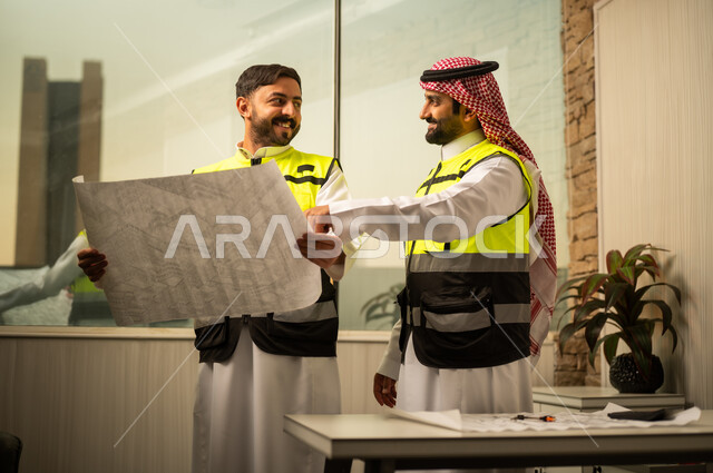 The concept of engineering planning, following project details, reviewing the construction plan, a Saudi Gulf Arab engineer wearing a safety vest standing and holding an engineering plan while following the explanation alongside a colleague, the use of paper plans in engineering work, a modern work environment in the construction sector, the concept of urban development and project management.