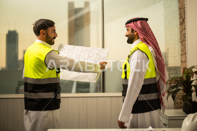 متابعة تفاصيل المشروع، مراجعة مخطط البناء، مفهوم التخطيط الهندسي، مهندس عربي خليجي سعودي يرتدي سترة السلامة يقف ويمسك مخطط هندسي يتابع الشرح برفقة زميلة، استخدام المخططات الورقية في العمل الهندسي، بيئة عمل حديثة في قطاع الإنشاءات، مفهوم التطوير العمراني وإدارة المشاريع