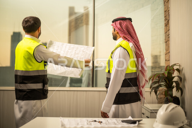 مراجعة مخطط البناء، متابعة تفاصيل المشروع، مفهوم التخطيط الهندسي، مهندس عربي خليجي سعودي يرتدي سترة السلامة يقف ويمسك مخطط هندسي يتابع الشرح برفقة زميلة، استخدام المخططات الورقية في العمل الهندسي، بيئة عمل حديثة في قطاع الإنشاءات، مفهوم التطوير العمراني وإدارة المشاريع