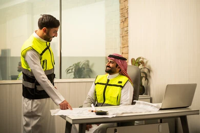 Monitoring the progress of the project, discussing work plans, professional collaboration within the engineering office, the concept of project management, planning and organizing in the work environment, a Saudi Gulf Arab engineer sitting on an office chair discussing work details with a colleague, using a laptop in engineering work, a modern work environment in the construction sector.