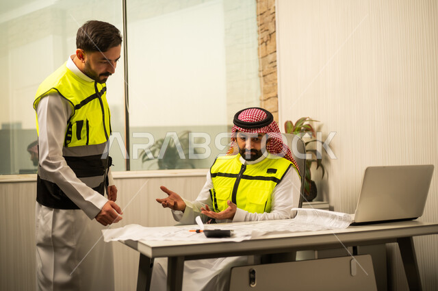 مناقشة مخططات العمل، التعاون المهني داخل المكتب الهندسي، متابعة سير المشروع، مفهوم إدارة المشاريع، التخطيط والتنظيم في بيئة العمل، مهندس عربي خليجي سعودي يجلس على كرسي مكتبي يناقش تفاصيل العمل برفقة زميلة، استخدام الحاسب المحمول في الأعمال الهندسية، بيئة عمل حديثة في قطاع الإنشاءات