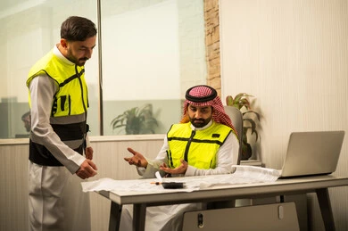 Discussion of work plans, professional collaboration within the engineering office, monitoring project progress, the concept of project management, planning and organization in the work environment, a Saudi Gulf Arab engineer sitting on an office chair discussing work details with a colleague, using a laptop in engineering work, a modern work environment in the construction sector.