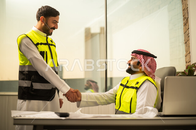 التعاون المهني داخل المكتب الهندسي، مفهوم الشراكة في بيئة العمل، إتمام اتفاق عمل بين الزملاء، الثقة والاحترام المتبادل في العمل، مهندس عربي خليجي سعودي يرتدي سترة السلامة يقف ويصافح زميله، استخدام التكنولوجيا في إدارة المشاريع، بيئة عمل حديثة في قطاع الإنشاءات