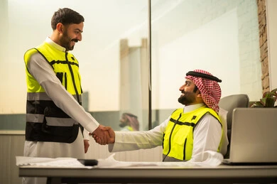 Professional collaboration within the engineering office, the concept of partnership in the work environment, completing a work agreement between colleagues, mutual trust and respect at work, a Saudi Gulf Arab engineer wearing a safety vest standing and shaking hands with his colleague, using technology in project management, a modern work environment in the construction sector.