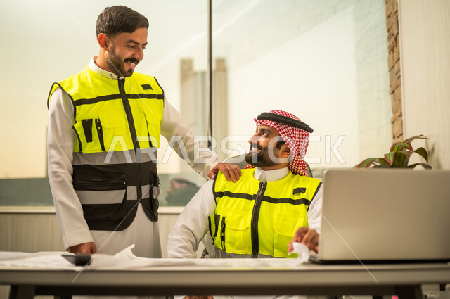 استخدام التكنولوجيا في التخطيط الهندسي، إدارة المشاريع الهندسية، مفهوم العمل الجماعي بين المهندسين، مهندس عربي خليجي سعودي يرتدي الشماغ وسترة أمان يعمل على جهاز كمبيوتر محمول برفقة زميله