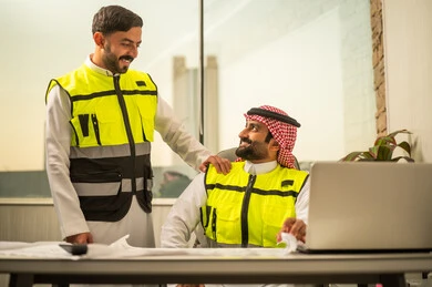 استخدام التكنولوجيا في التخطيط الهندسي، إدارة المشاريع الهندسية، مفهوم العمل الجماعي بين المهندسين، مهندس عربي خليجي سعودي يرتدي الشماغ وسترة أمان يعمل على جهاز كمبيوتر محمول برفقة زميله