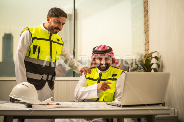 مفهوم العمل الجماعي بين المهندسين، إدارة المشاريع الهندسية، استخدام التكنولوجيا في التخطيط الهندسي، مهندس عربي خليجي سعودي يرتدي الشماغ وسترة أمان يعمل على جهاز كمبيوتر محمول برفقة زميله