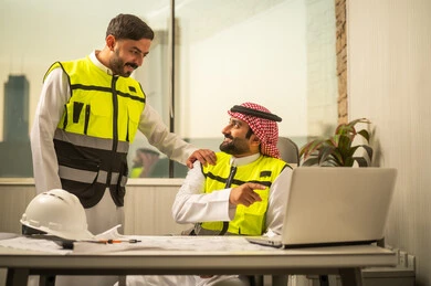 إدارة المشاريع الهندسية، مفهوم العمل الجماعي بين المهندسين، استخدام التكنولوجيا في التخطيط الهندسي، مهندس عربي خليجي سعودي يرتدي الشماغ وسترة أمان يعمل على جهاز كمبيوتر محمول برفقة زميله