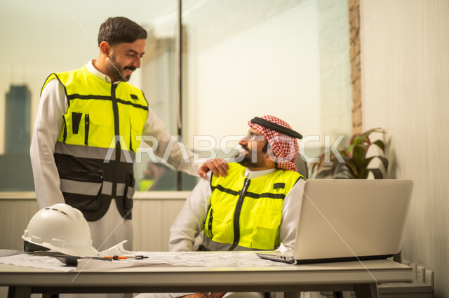 Teamwork in engineering projects, the concept of cooperation and coordination among colleagues, monitoring the workflow within an engineering office, two Arab Gulf engineers from Saudi Arabia wearing safety vests are discussing.