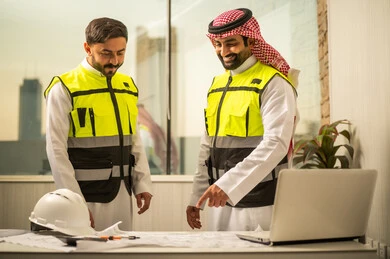 التخطيط الهندسي وإدارة المشاريع, مفهوم السلامة المهنية في بيئة العمل, مراجعة المخططات الهندسية قبل التنفيذ, مهندسان عربيان خليجيان سعوديان يرتديان سترات السلامة يقفان أمام مكتب عمل يتفقدان الرسومات