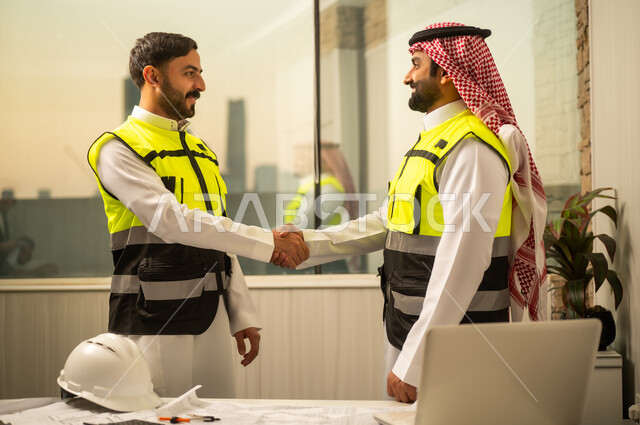 إدارة المشاريع الهندسية, مفهوم التواصل وبناء الثقة في بيئة العمل, متابعة سير العمل, مهندسان عربيان خليجيان سعوديان يرتديان سترات السلامة يتبادلان المصافحة داخل المكتب