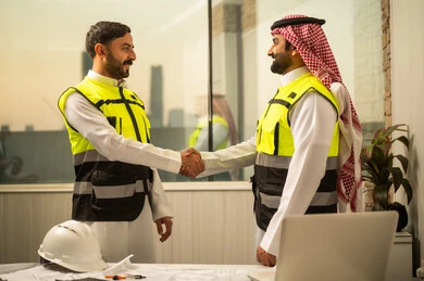 إدارة المشاريع الهندسية, مفهوم التواصل وبناء الثقة في بيئة العمل, متابعة سير العمل, مهندسان عربيان خليجيان سعوديان يرتديان سترات السلامة يتبادلان المصافحة داخل المكتب
