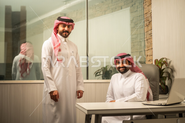 مفهوم التعاون الوظيفي داخل الشركات، استخدام التكنولوجيا في متابعة الأعمال، موظف عربي خليجي سعودي  يرتدي الثوب الابيض و الشماغ يجلس أمام الحاسب المحمول برفقة زميلة، النظر للكاميرا بتعابير السعادة و السرور