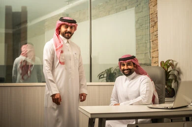 مفهوم التعاون الوظيفي داخل الشركات، استخدام التكنولوجيا في متابعة الأعمال، موظف عربي خليجي سعودي  يرتدي الثوب الابيض و الشماغ يجلس أمام الحاسب المحمول برفقة زميلة، النظر للكاميرا بتعابير السعادة و السرور