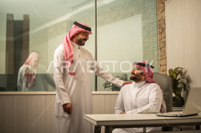 مفهوم الإدارة والتنظيم، استخدام الأجهزة الذكية في الأعمال المكتبية، موظف عربي خليجي سعودي يرتدي الثوب الابيض و الشماغ يجلس امام الحاسب المحمول يناقش العمل مع زميله