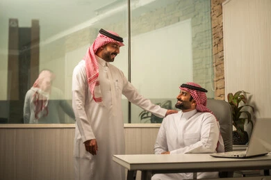 مفهوم الإدارة والتنظيم، استخدام الأجهزة الذكية في الأعمال المكتبية، موظف عربي خليجي سعودي يرتدي الثوب الابيض و الشماغ يجلس امام الحاسب المحمول يناقش العمل مع زميله