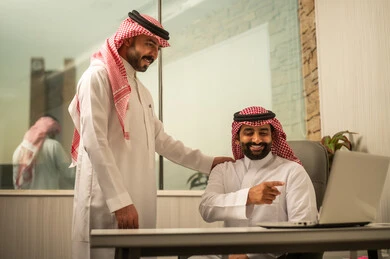 مناقشة فكرة داخل بيئة عمل مكتبية، متابعة مهمة عمل عبر حاسب محمول، مفهوم التعاون المهني، توجيه وإرشاد أثناء العمل، موظف عربي خليجي سعودي يرتدي الثوب و الشماغ يجلس أمام حاسب محمول  يتحدث مع زميلة ويشير إلى الشاشة، أجواء عمل إيجابية، بيئة مكتبية عصرية