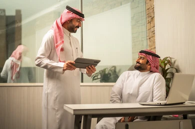 استخدام التقنية الحديثة في بيئة العمل، تبادل فكرة ومعلومة داخل المكتب، متابعة سير العمل باستخدام جهاز لوحي، مفهوم التواصل المهني، موظف عربي خليجي سعودي يقف بجانب المكتب يمسك جهاز لوحي يقوم بمناقشة زميلة، بيئة عمل مكتبية عصرية